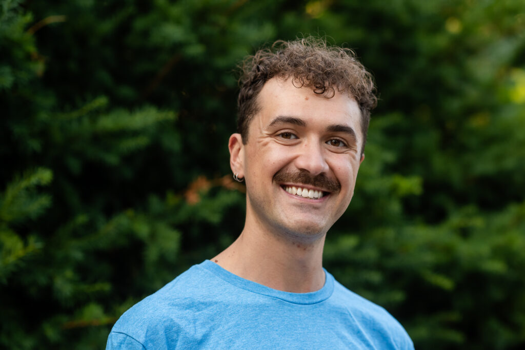 The image shows Jeff, a white man with brown, curly hair and moustache, smiling at the camera. He wears a blue shirt and stands against a background of dark green trees